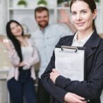 A smiling businesswoman holding a home insurance policy with clients in the background.